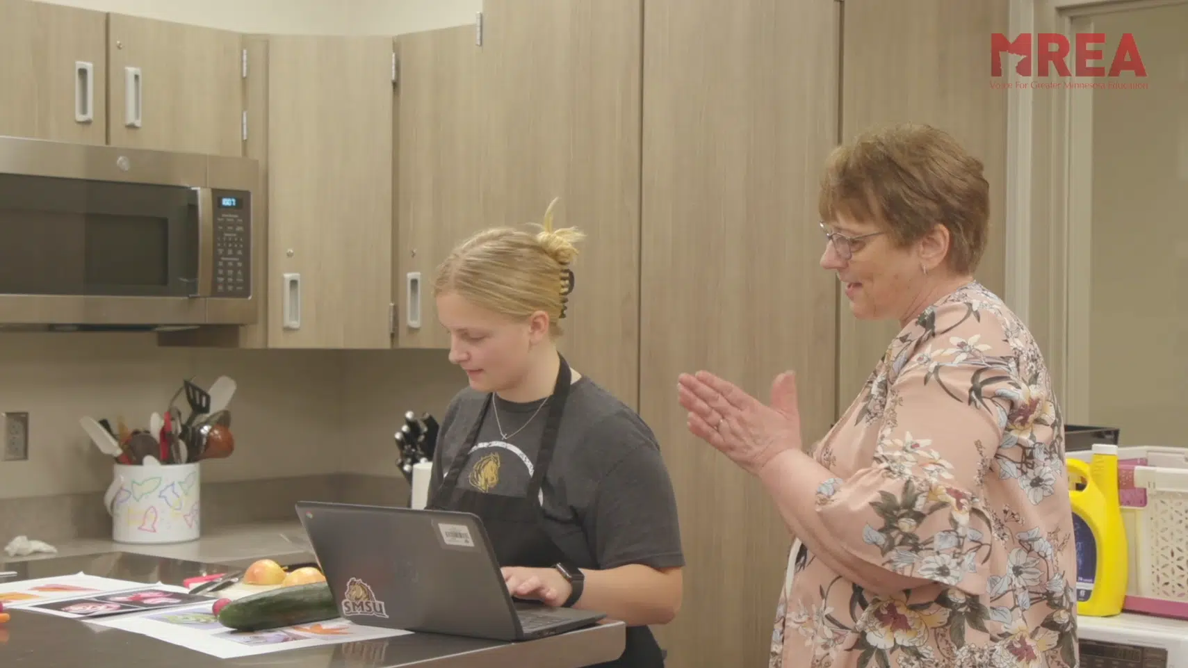A teacher in a Family and Consumer Science class kitchen smiling and talking to a student who is working at the counter, wearing an apron. There is a laptop on the counter.