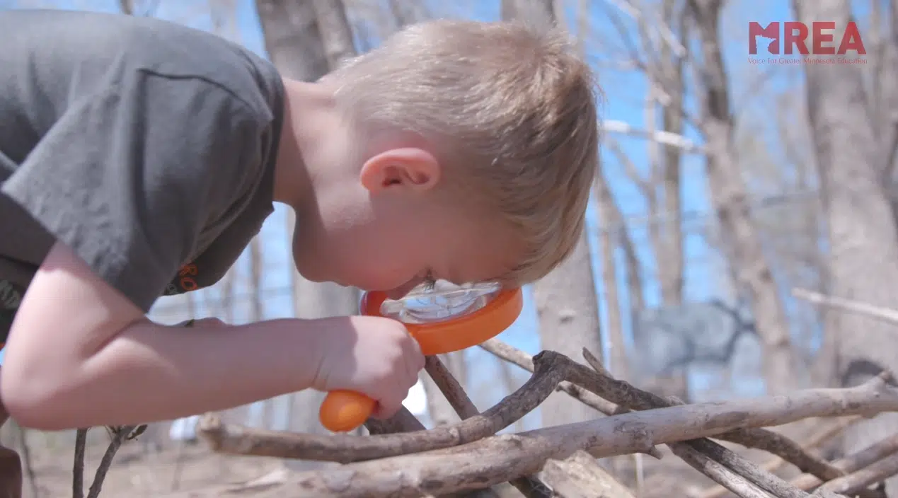 An elementary student using an orange magnifying glass to inspect the surface of a fallen branch.