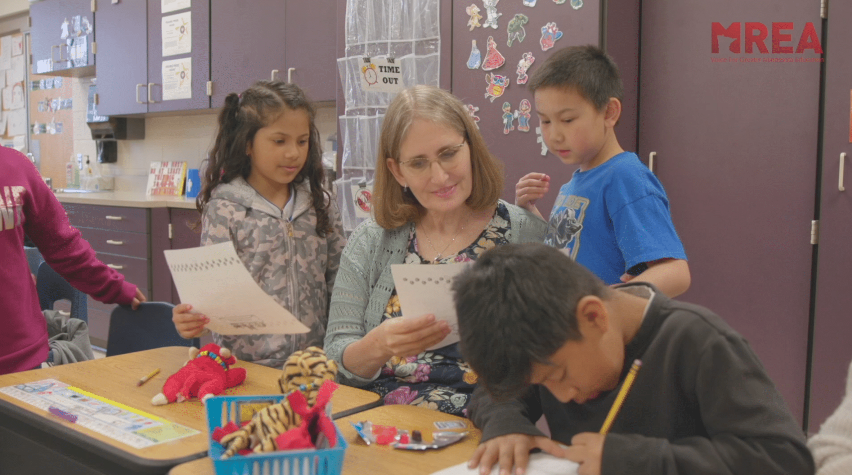 A teacher in a classroom, encircled by students, reads a worksheet while the students look on