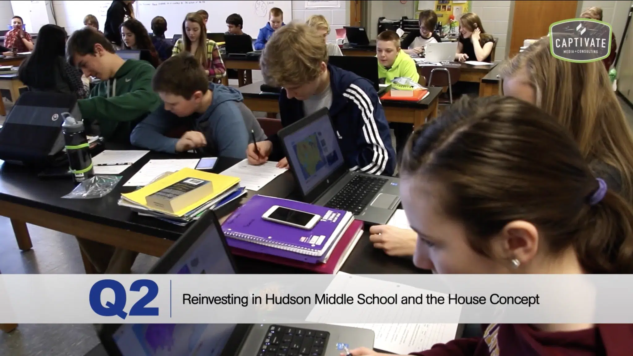 A screenshot from the video. A classroom of students working at tables.
