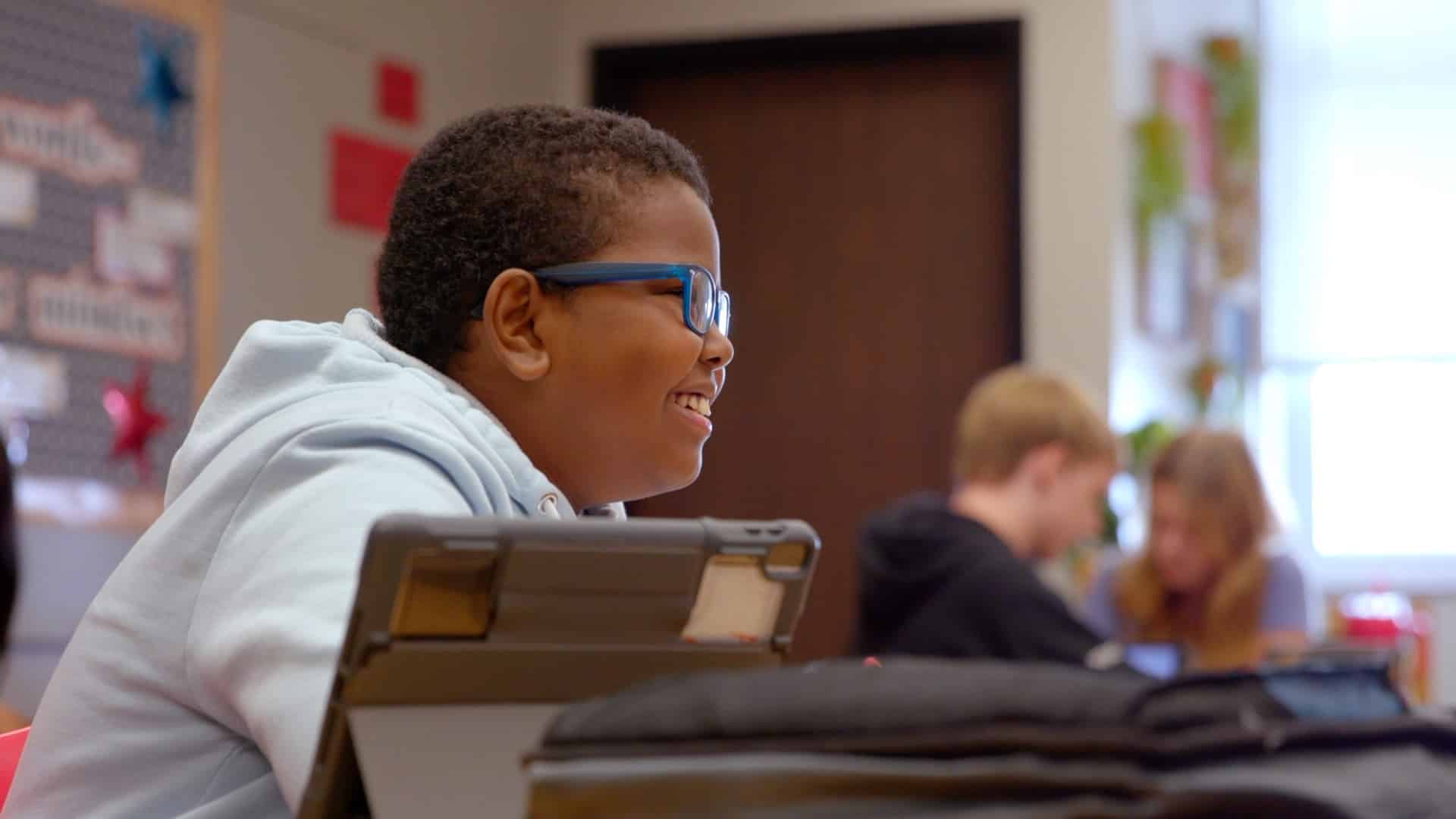 student smiling, wearing glasses, with an iPad on his desk