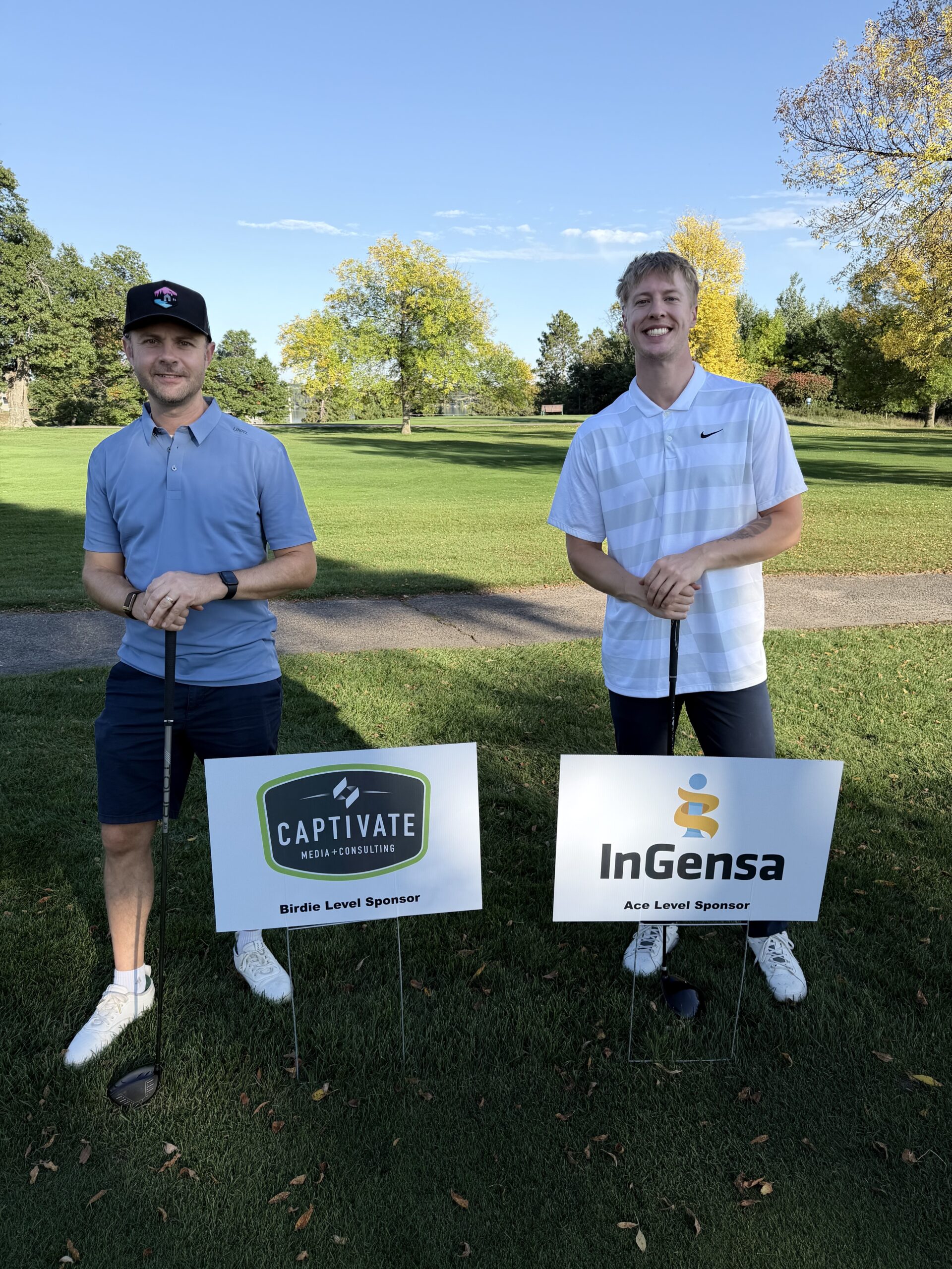 two men leaning on golf clubs standing behind small signs with company logos on them