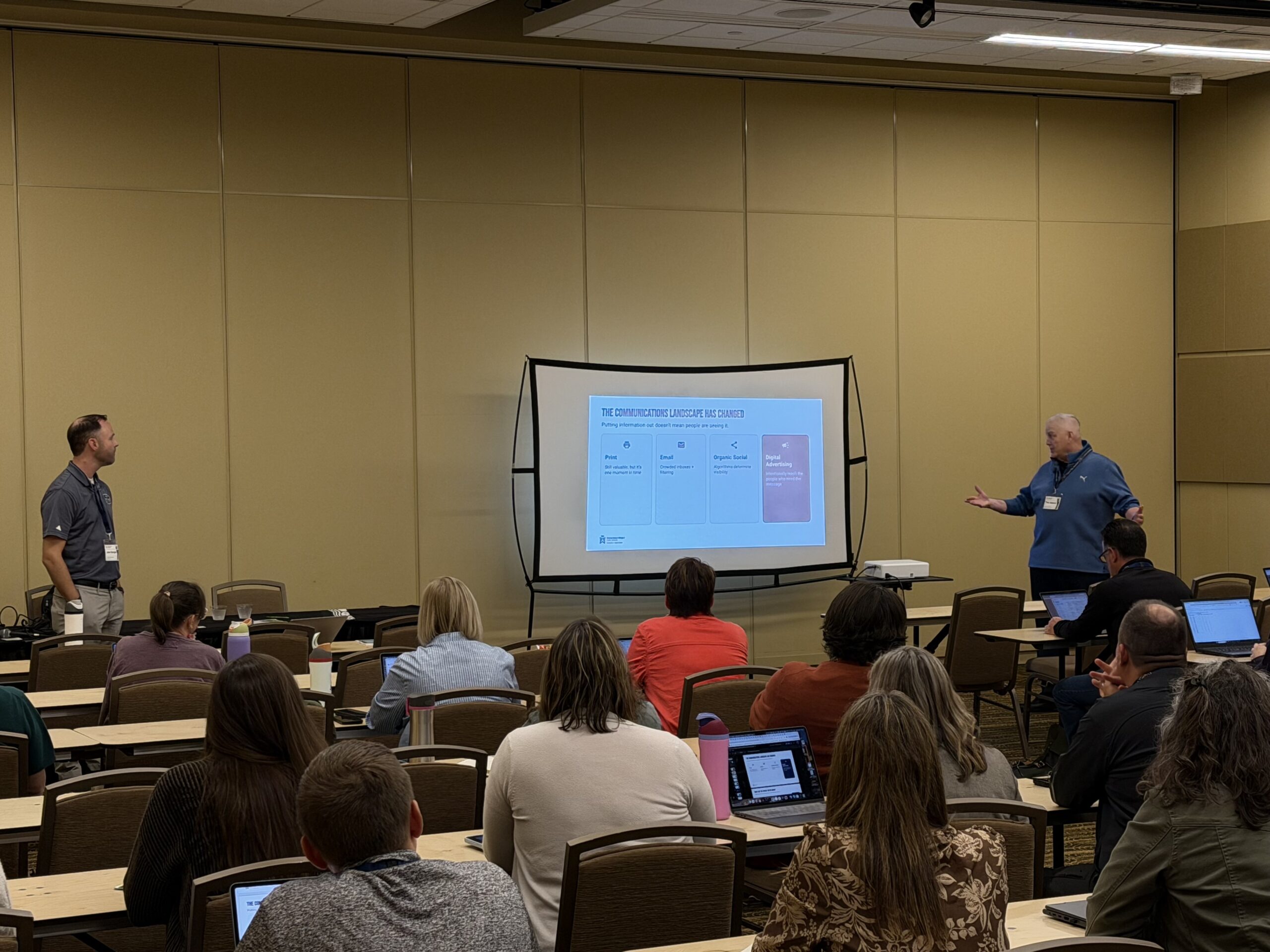 Two men presenting on either side of a screen in front of a crowd