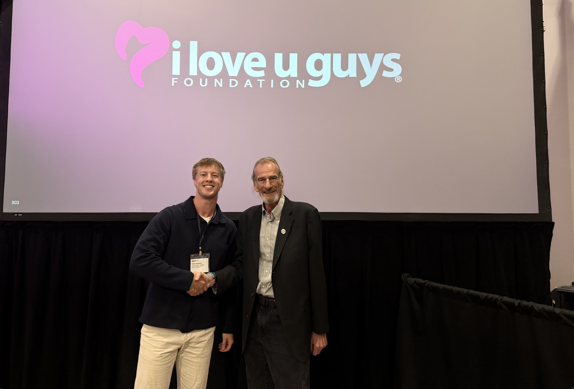 Two men smiling and shaking hands in front of a sign that says "I love u guys"